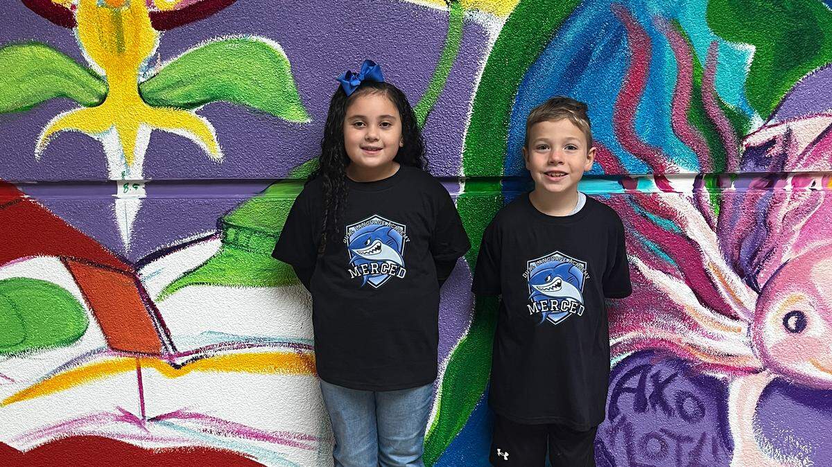 Aliviya, a first grader, and Rhys, a second grader are standing in front of a brightly colored mural wearing matching shirts in support of the Dual Language Academy.