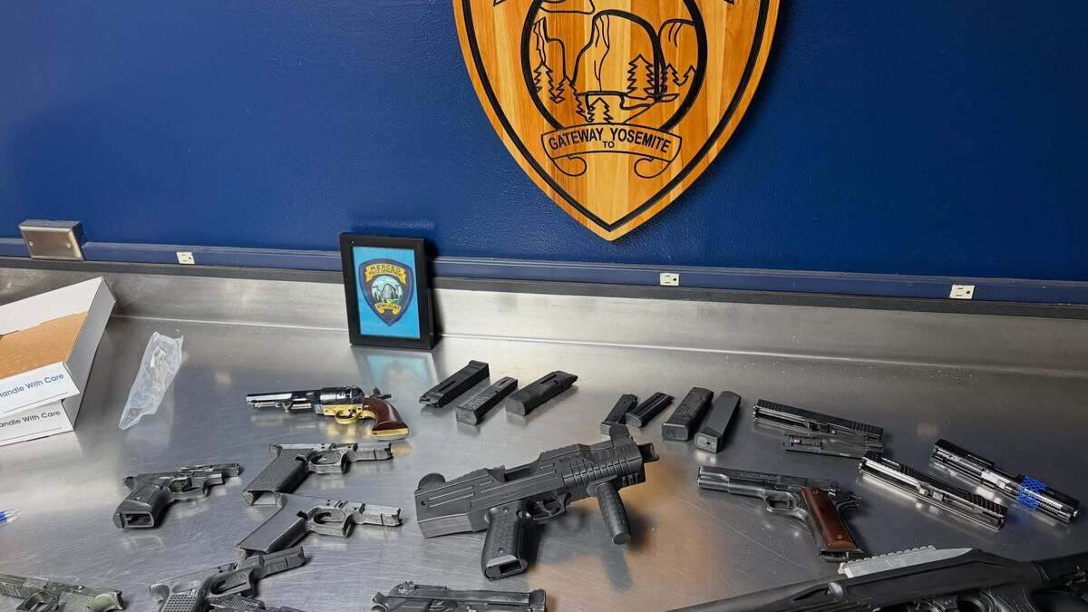 An assortment of 11 guns, including an automatic weapon spread on a table under the Merced Police Department crest.
