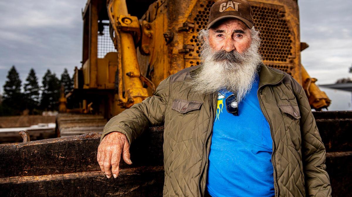 Gary Reneiro, 83, leans against piece of heavy machinery he uses to move earth for various jobs in Merced, Calif., on Thursday, Dec. 7, 2023.