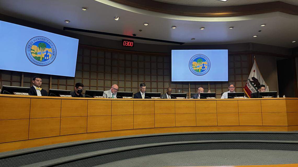Eight people sitting behind a wooden dais, behind them screens display the logo of the City of Merced.