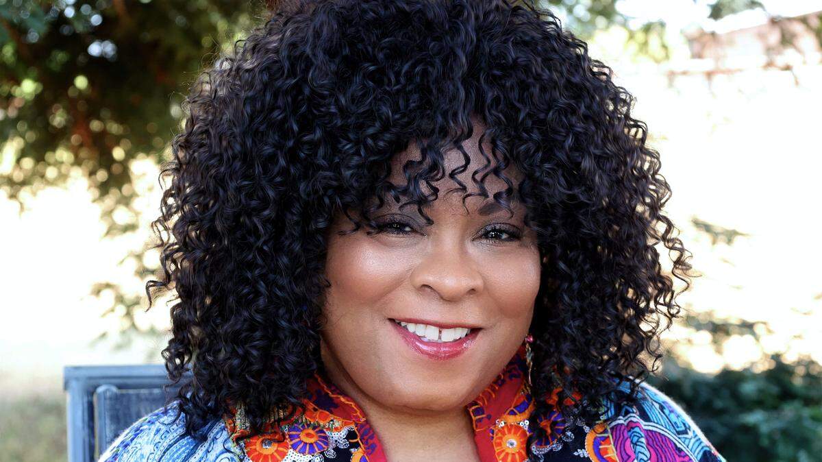 McMillon faces the camera, she is wearing a bright top and wearing her curly hair down.