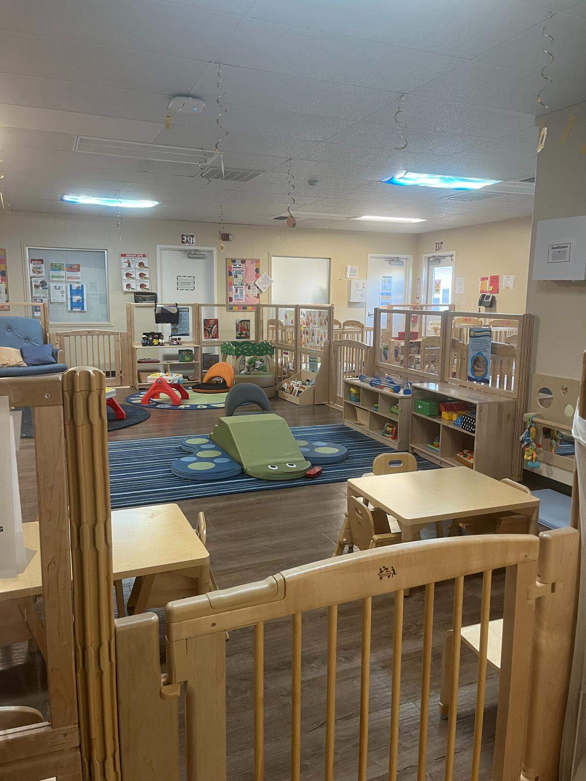 A room sectioned off by wooden babygates, one section has play equipment for infants and toddlers and the other is filled with cribs.