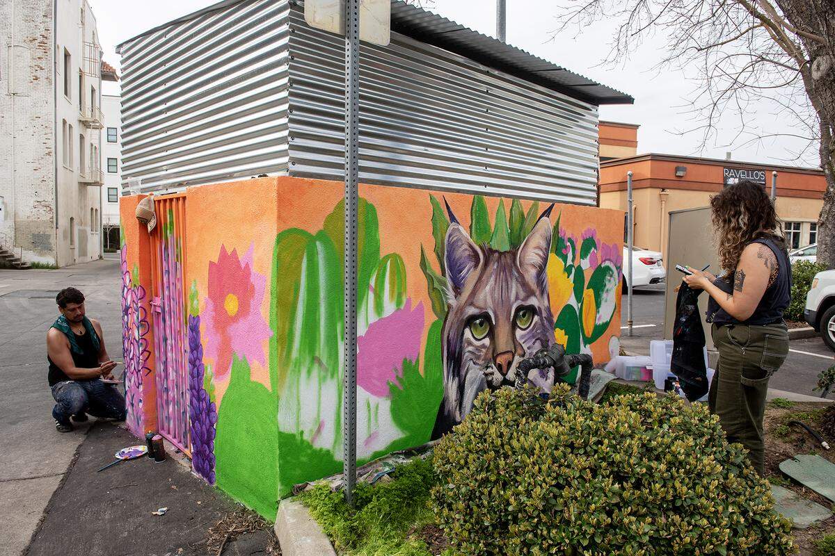 Fresno artist Andrea Torres, 35, right, paints a mural she created while fellow Fresno artist Mauro Carrera, 38, left, assists her with the painting of a flower, on the walls of a dumpster enclosure in a parking lot off of West 18th Street in Merced, Calif., on Feb. 27, 2025. Torres said her mural is meant to represent community pride while including elements of the natural valley landscape and touching on the connection the city has with nearby UC Merced.