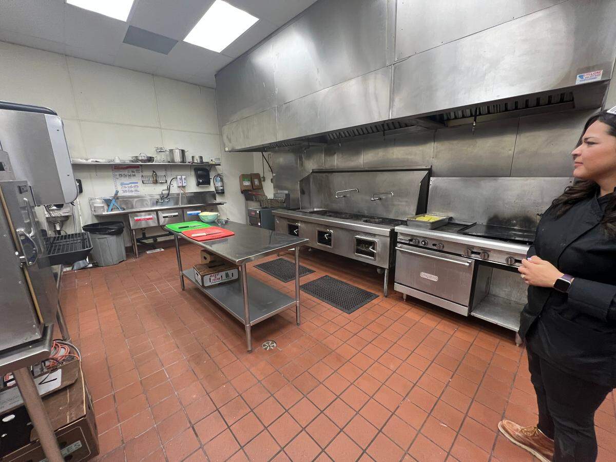 A woman in a chef coat stands in a commercial kitchen. 