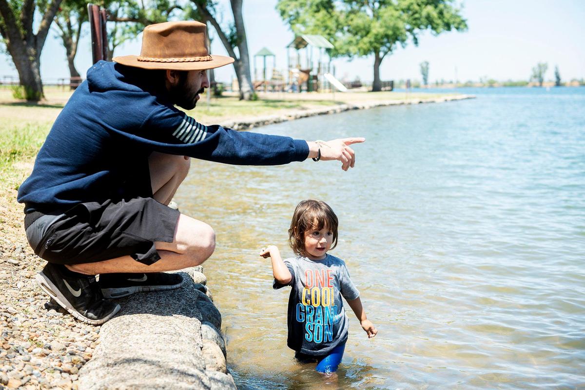 Fernando Arias, 22, of Merced, left, looks on as son Celeb, 2, right, throws rocks while cooling off at Lake Yosemite in Merced, Calif., on Thursday, July 8, 2021. According to Arias, he and other family members went to the lake to cool off and barbecue.