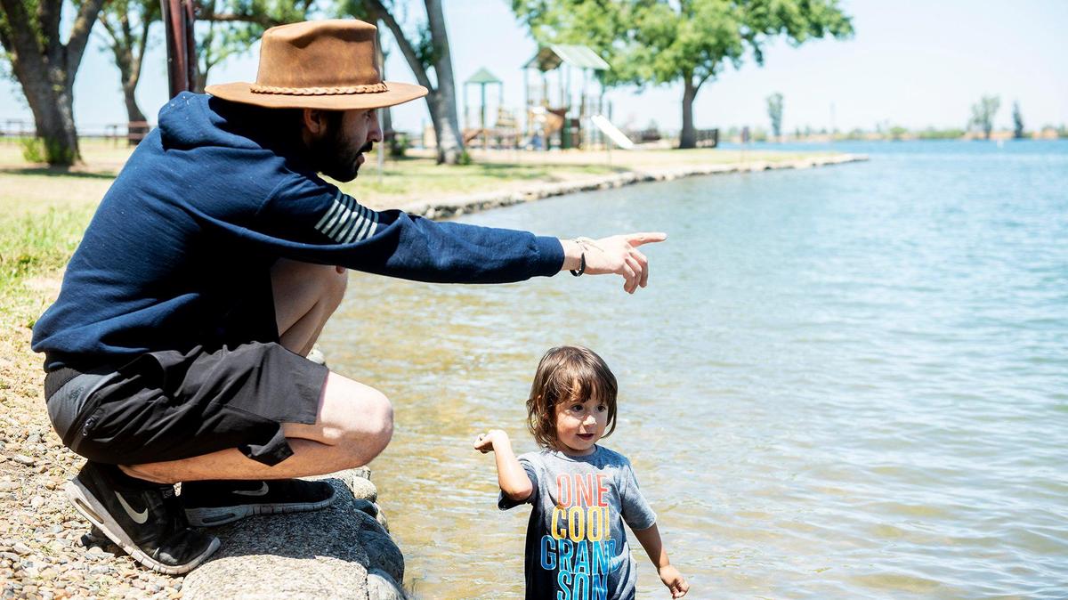 Fernando Arias, 22, of Merced, left, looks on as son Celeb, 2, right, throws rocks while cooling off at Lake Yosemite in Merced, Calif., on Thursday, July 8, 2021. According to Arias, he and other family members went to the lake to cool off and barbecue.