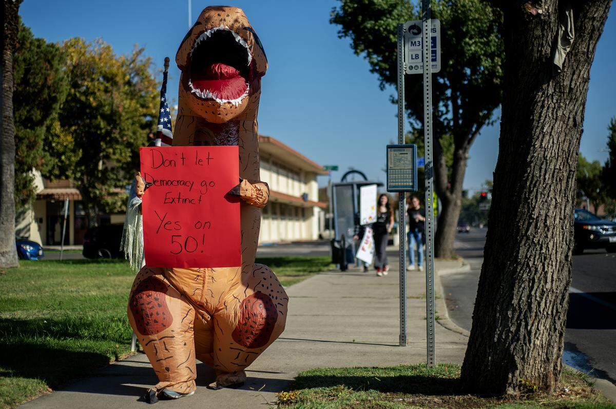 A person in a dinosaur costume carries a sign along M Street during a “No Kings” protest in Merced, Calif., on Saturday, Oct. 18, 2025. 