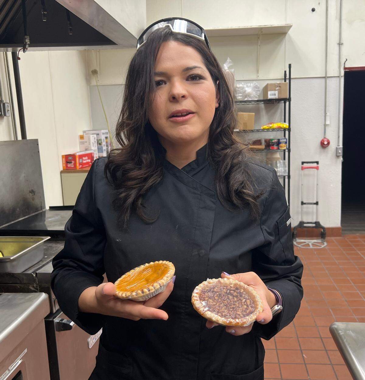 A woman in a chef coat, holds a miniature pecan pie and a miniature sweet potato pie. 