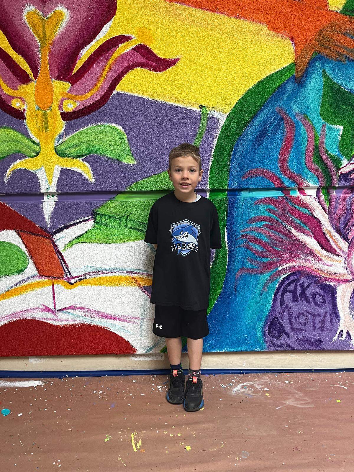 Second-grade student Rhys stands in front of a mural painted by Jazz Diaz, a local artist and DLA parent.