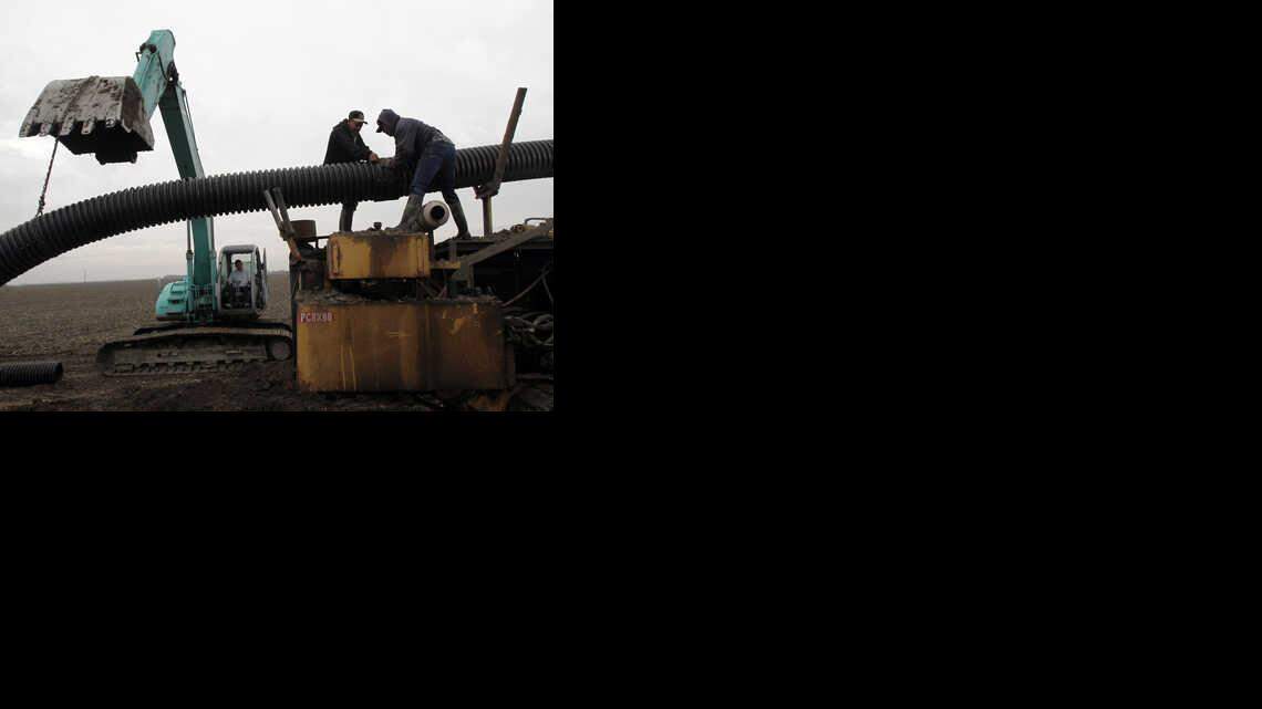 Rene Ruiz, left, waits in an excavator as Julian Flores, center and Alfonso Velasquez couple two large corrugated perforated plastic pipe sections as part of a drainage system.