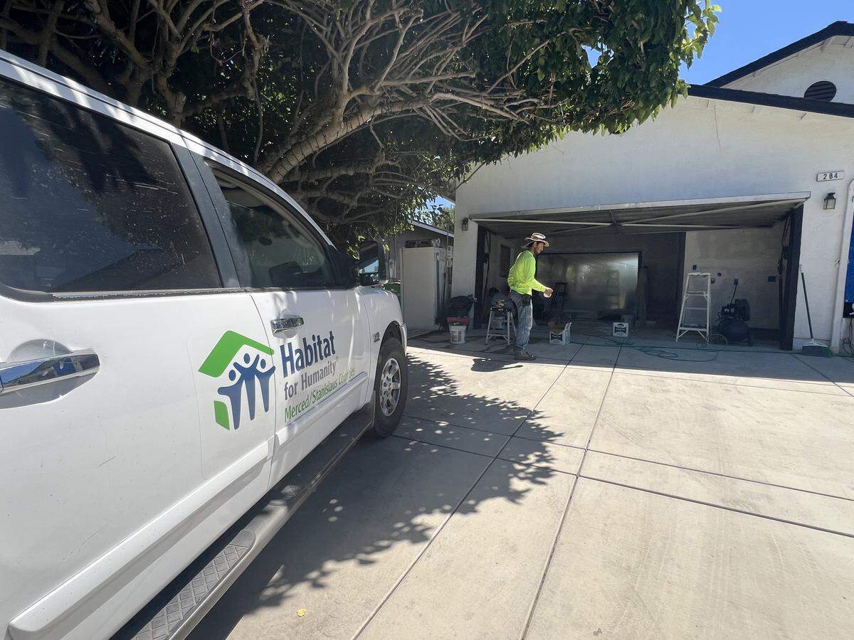 A white truck with the Habitat for Humanity logo on its side parks in the driveway of a house where men work on the garage walls.
