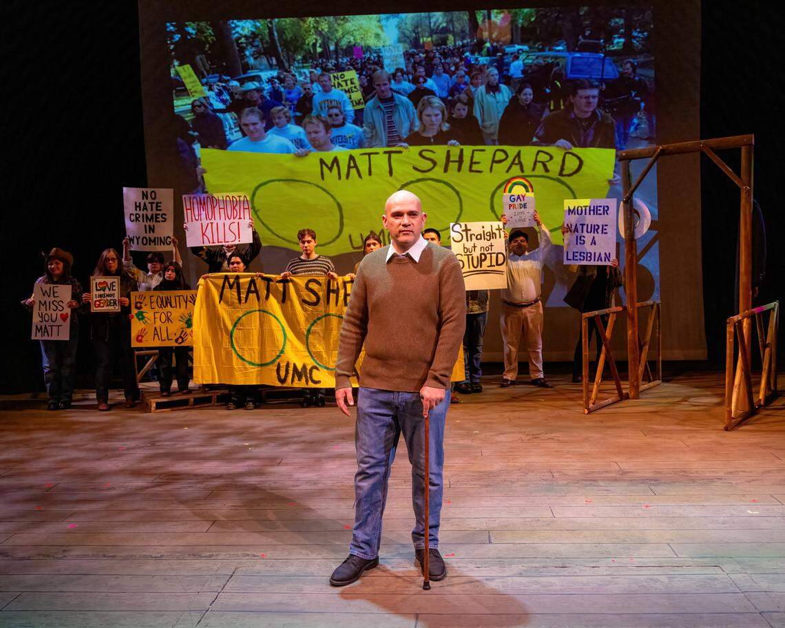 Carlo Martinez onstage during a rehearsal of The Laramie Project, he is holding a cane. Behind him are actors dressed as protesters as well as real photos of the protesters in Laramie after Matthew Shepard’s murder.