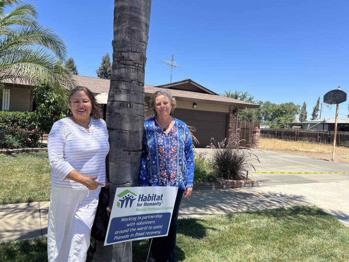 Two women stand next to a lawn sign in front of a home where the driveway is wet concrete.