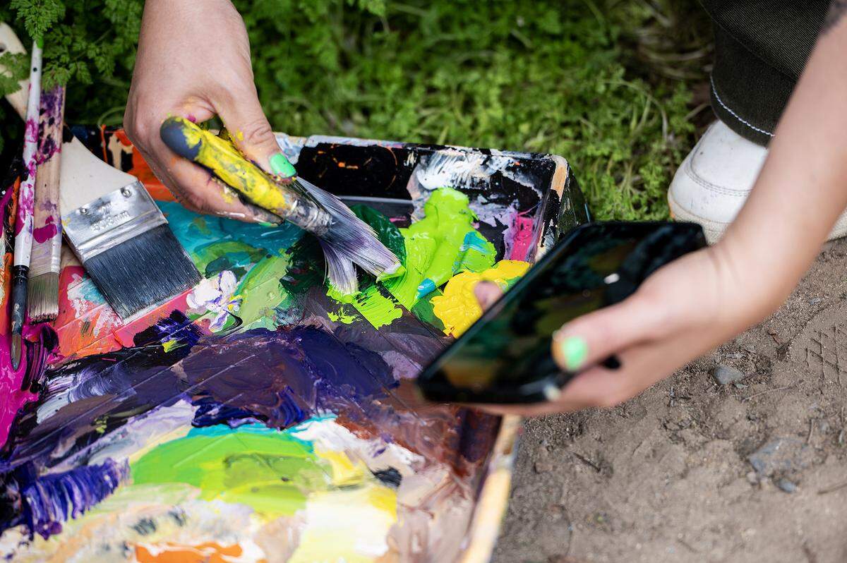 Fresno artist Andrea Torres, 35, dips her brush into paint while referencing her mural design on her phone as she paints the walls of a dumpster enclosure in a parking lot off of West 18th Street in Merced, Calif., on Feb. 27, 2025. Torres said her mural is meant to represent community pride while including elements of the natural valley landscape and touching on the connection the city has with nearby UC Merced.
