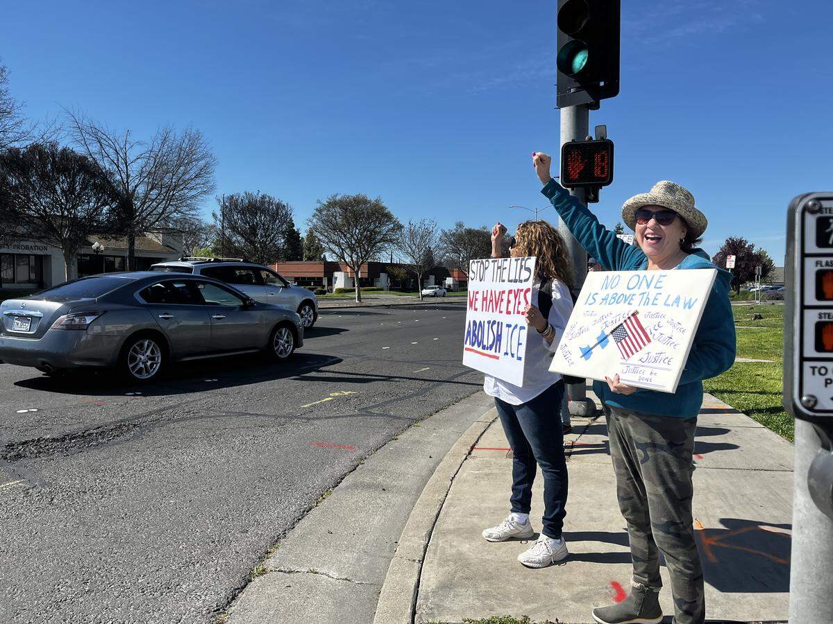 Protestors attend a demonstration against the U.S. war with Iran. Two women are standing on a corner, holding their fists in the air.