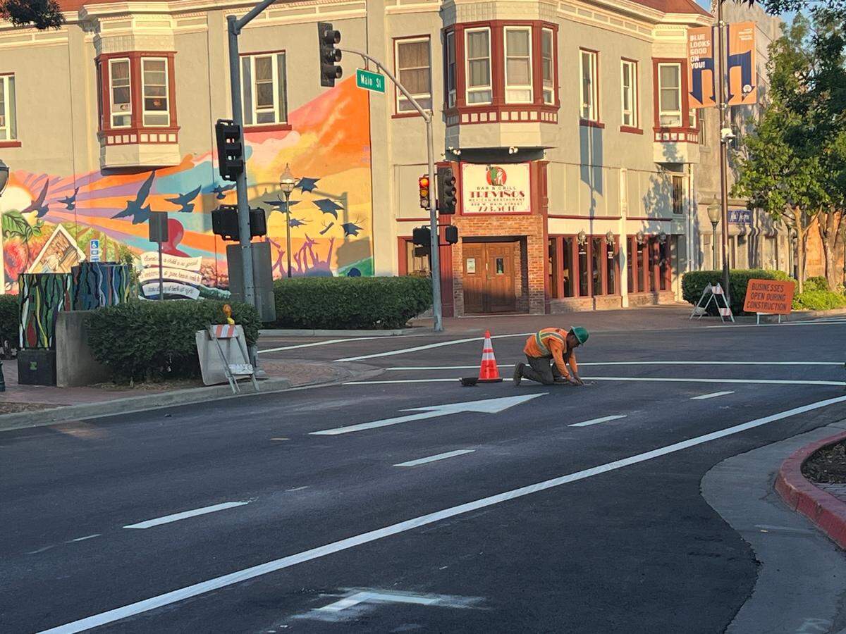 A man in a orange vest kneels in the middle of an empty road and looks at something in the road. 