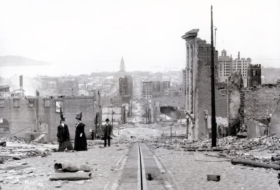 Black and white photo of three people in period attire walking down a street in which buildings are destroyed.
